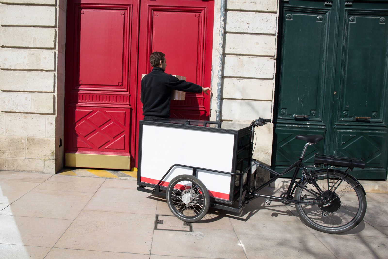 Ein Mann ist in schwarz gekleidet und steht vor einer roten Tür. Hinter ihm steht ein Lastenrad mit einer großen weißen Kiste vor dem Lenkrad.